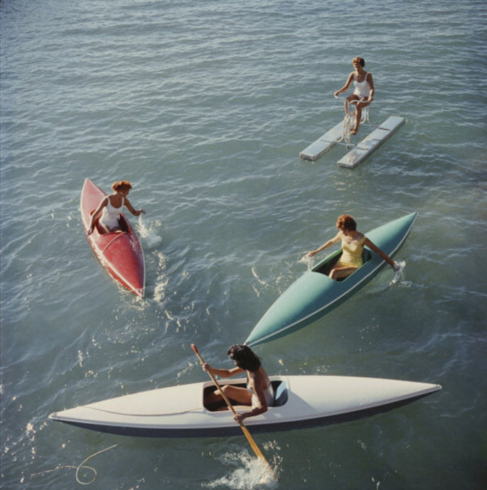 Lake Tahoe Trip by Slim Aarons - Photograph overlooking a green coloured Lake Tahoe with 1950's women bathers canoeing and enjoying a day on the water.