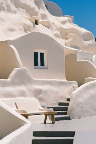 Mystique Hotel by Via Tolila - An iconic Santorini view of white stone buildings carved into the rugged Caldera cliffs, surrounded blue sky and blue water.