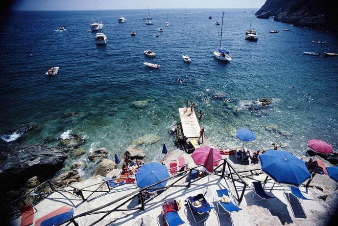 Porto Ercole by Slim Aarons - A vintage photograph of a coastal scene with beach umbrellas and boats moored alongside the shore.