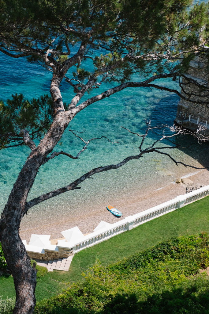 Private Beach by Via Tolila - A photograph of a pristine beach with white umbrellas in the distance and lush green oceanfront gardens. 