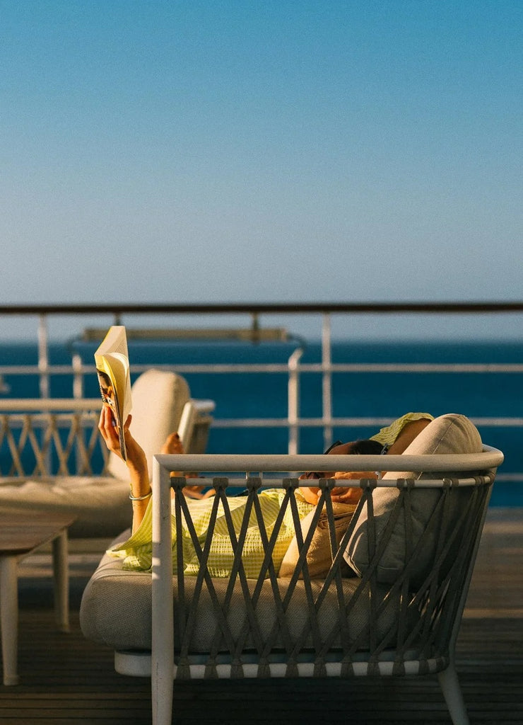Sunset Hour by Via Tolila - A photographic print of a person reading a book on a chair by the sea with a clear blue sky.