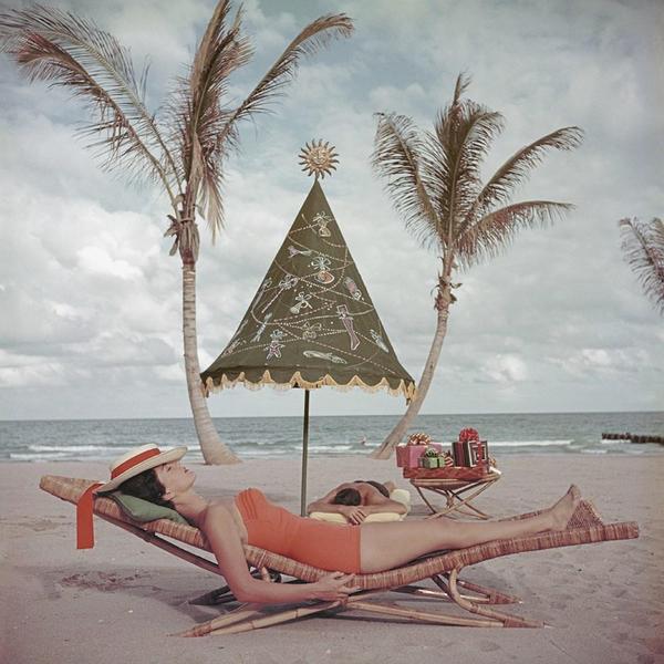 Palm Beach Idyll by Slim Aarons - A photograph captured in 1955 of a young lady sunbathing in a one piece orange swimsuit on a lounge on the beach. 