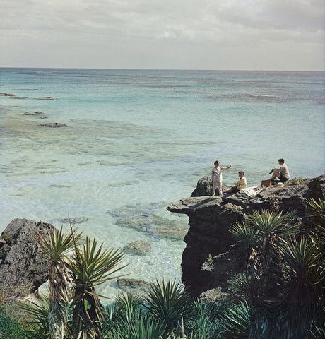 A Nice Spot for Lunch by Slim Aarons - A group of three have a picnic at a rocky coastline besides the ocean.