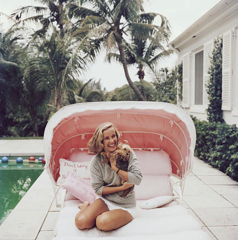 Alice Topping by Slim Aarons - A vintage photograph of lady laughing, cuddling her puppy on a pink outdoor daybed by the pool.