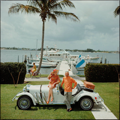 All Mine by Slim Aarons - A married couple standing in front of a white sports car and white boats moored in front of a lake.