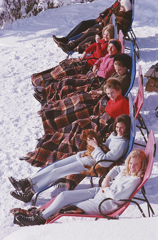 Après Ski by Slim Aarons - Skiers lounging in the snow, lined up in recliner ski chairs. One woman holds a dog.