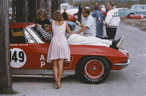 Bahamas Speed Week I by Slim Aarons - A man sitting on a car having a conversation with a woman standing, at a car racing event.