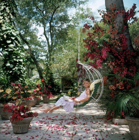 Barbados Bliss by Slim Aarons - Lady sits in a white swing chair surrounded by colourful bougainvillaea in a stunning garden setting