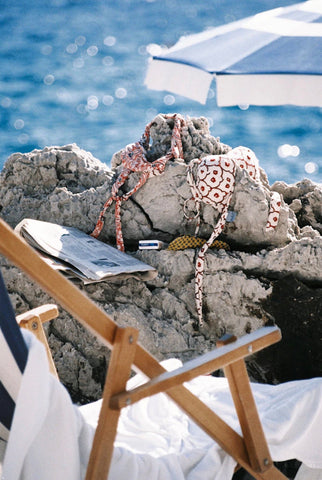 Beach Equipped by Akila Berjaoui. A film photograph of a beach scene with blue striped sun umbrella, deck chair, newspaper and bikini tops drying on the rocks.