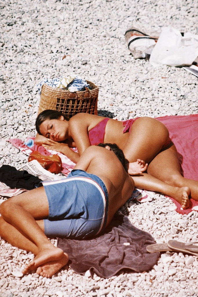 Capri Slumbers by Akila Berjaoui. Two bronze beachgoers sleep on the rocky beach of Capri. With a woven beach bag and newspaper nearby, the girl wears a hot pink bikini and the boy wears blue shorts.