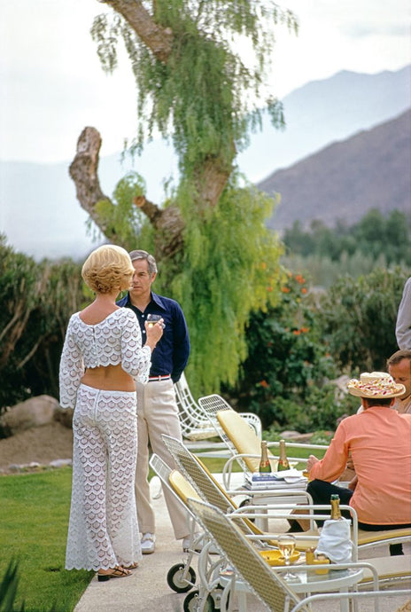Champagne by the Pool by Slim Aarons - A lady in glamorous crochet 70s outfit chats to a man in navy shirt with champagne glass in hand by the pool