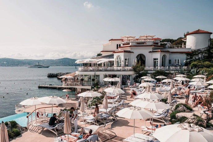 Eden Afternoons by Stuart Cantor - Hotel du Cap, iconic oceanfront luxury hotel, captured with holidaymakers relaxing beneath white sun umbrellas in the foreground with glittering ocean in the distance.