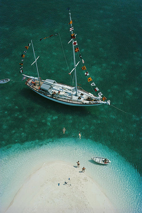 Exuma Holiday by Slim Aarons - A yacht with many flags beside a sandbank. 