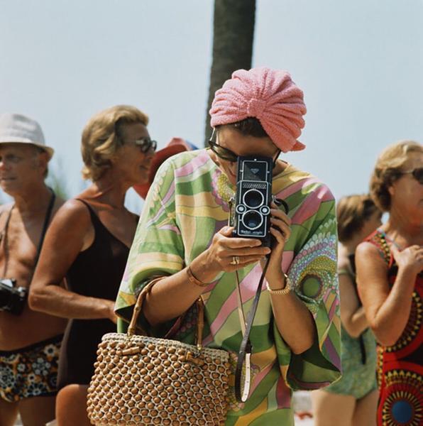 Grace Kelly Photographs by Getty Images (UK) Ltd - Princess Grace of Monaco taking a photograph. She wears a pink and green 70s style dress and pink hair wrap.