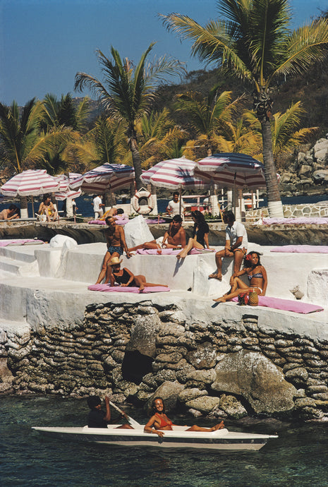 La Concha Clientele by Slim Aarons - 1972 photograph of holidaymakers relaxing on pink sun-lounges, striped pink umbrellas on the cliff-side beach resort. Lady relaxes in a boat in the deep blue ocean below.