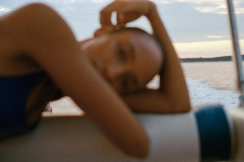 Looking Forward by Akila Berjaoui. An out of focus, film photograph of a girl close up. She is leaning to the side and the glittering water of the ocean is in the background. 