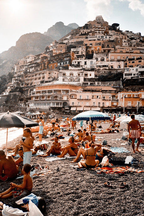 Positano by Akila Berjaoui - Captured on film, this landscape photograph features a sun-filled beach setting with beautiful buildings in the distance in terracotta tones