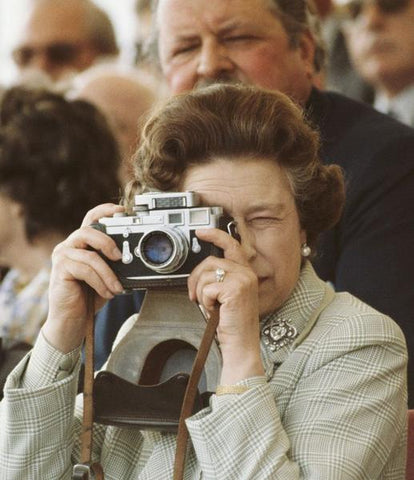 Queen Elizabeth Leica Camera by Getty Images (UK) Ltd - Queen Elizabeth II is taking a picture with her Leica M3 camera in 1982. She wears a plaid jacket, adorned with a brooch and is squinting into the camera.