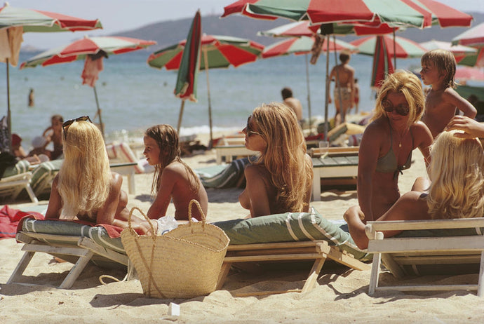 Saint-Tropez Beach by Slim Aarons - Sunbathers lounge on the beach with red and green striped beach umbrellas lining the backdrop.