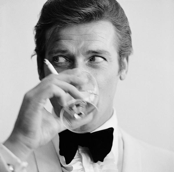 Shaken Not Stirred by Getty Images (UK) Ltd - This black and white photograph is a close up shot of Roger Moore taking a sip of a martini, with a cigarette in hand.