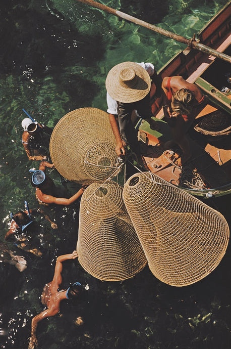 Snorkelling in The Shallows by Slim Aarons - A group of people snorkel with woven fishing nets off a boat in the shallow green water.