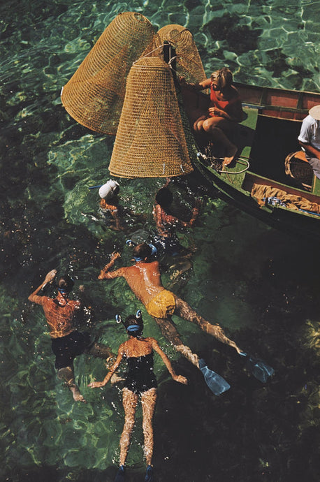 Snorkelling in Malta by Slim Aarons - A group of people snorkel with woven fishing nets off a boat in the shallow green water.