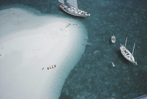 Stocking Island, Bahamas by Slim Aarons - Deep green ocean waters with a curved shoreline with white sand and white boats moored