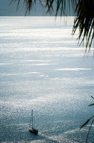 Summer Wine by Akila Berjaoui. A film photograph of a blue, glittering ocean, captured from a high vantage point. A sole yacht sails through the calm waters.