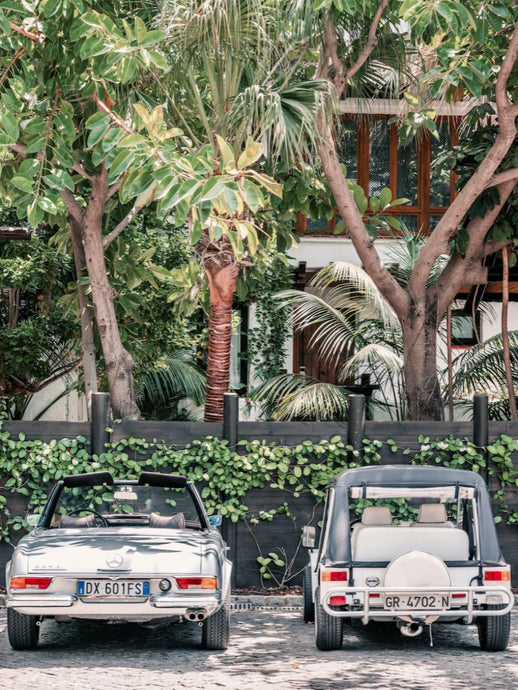 The Merc or the Moke? by Stuart Cantor - A photographic print of two retro cars parked by a hotel building with palm trees.