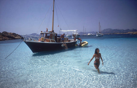  Yachting Trip by Slim Aarons - A lady wades ashore from a yacht in shallow, crystal blue waters.